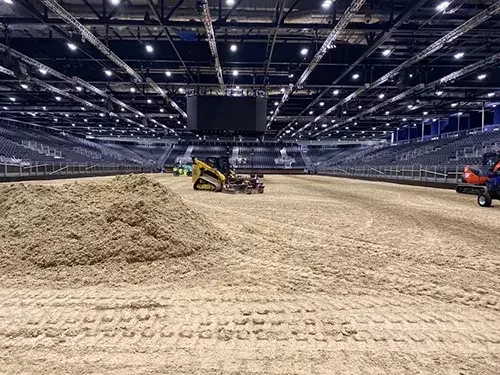 This image shows Total Equestrian Construction preparing the riding surface at the ExCeL London International Horse Show, with specialist equipment levelling sand footing inside the arena.