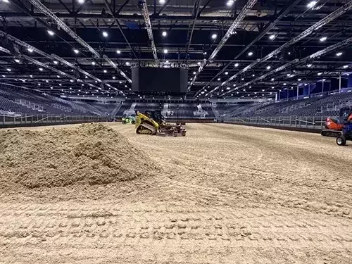 This image shows Total Equestrian Construction preparing the riding surface at the ExCeL London International Horse Show, with specialist equipment levelling sand footing inside the arena.