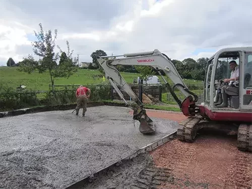 This image shows groundwork construction for an equestrian facility, with concrete being laid and levelled using a digger and manual tools.