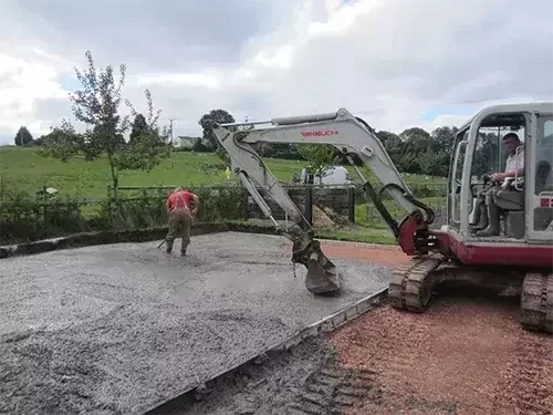This image shows groundwork construction for an equestrian facility, with concrete being laid and levelled using a digger and manual tools.