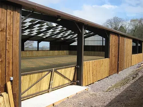 This image shows an indoor equestrian arena with timber cladding, skylights, and wooden kickboards around the riding area.