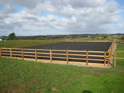 This image shows an outdoor equestrian arena with a rubber riding surface, surrounded by timber fencing, set in open countryside.