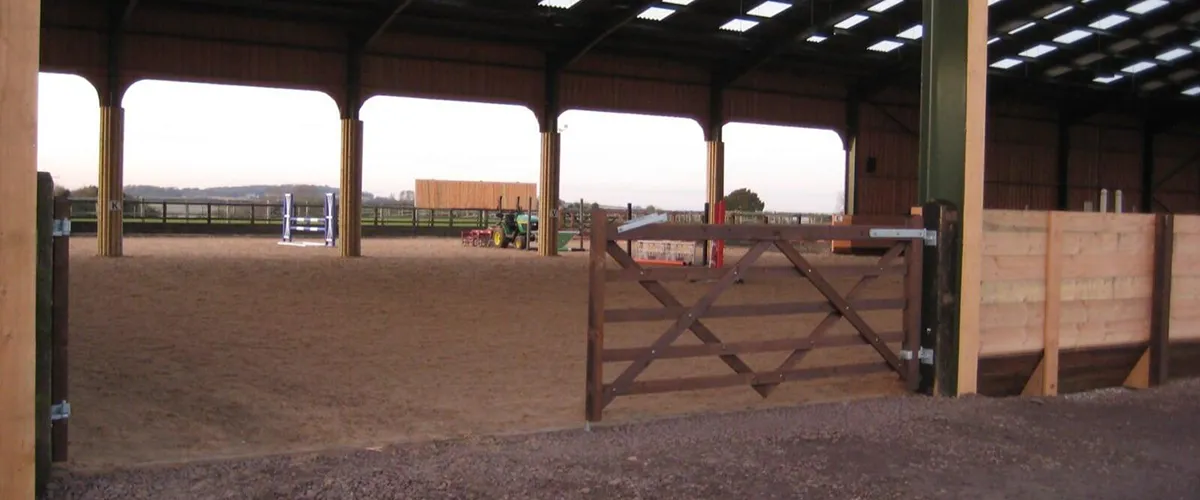 This image shows a covered outdoor equestrian arena with sand surface, timber fencing, and secure gated access for training and events.